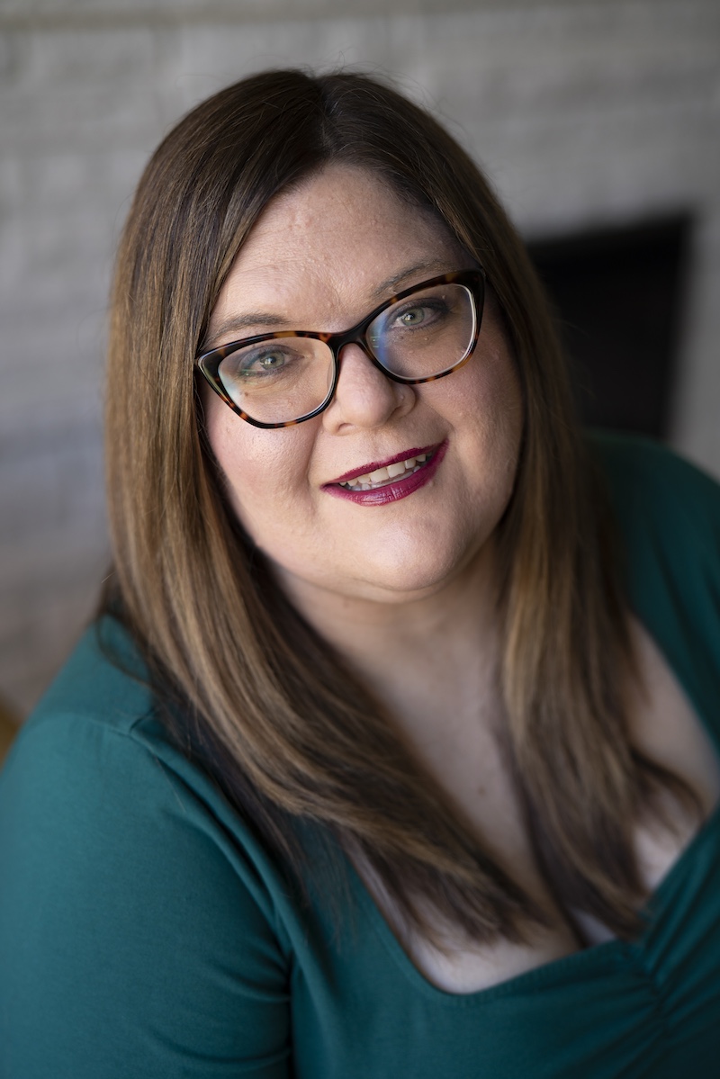 Becca Syme in front of a grey brick wall. She is wearing a green shirt and smiling.