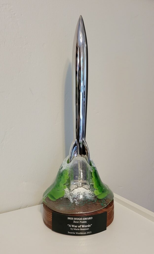 The Hugo trophy for the new speculative poetry award in the shape of a silver rocket ship with the following inscription: 2025 Hugo Award - Best Poem - "A War of Words" - by Marie Brennan - Seattle Worldcon 2025
